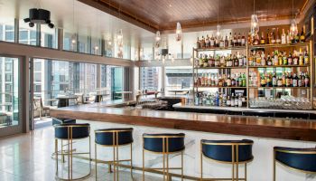 A modern hotel bar with a long counter, shelves of liquor bottles, hanging pendant lights, and several high stools by large windows.