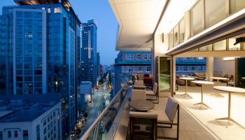 A modern high-rise balcony overlooks a downtown street lined with blue glass office buildings under a clear sky.
