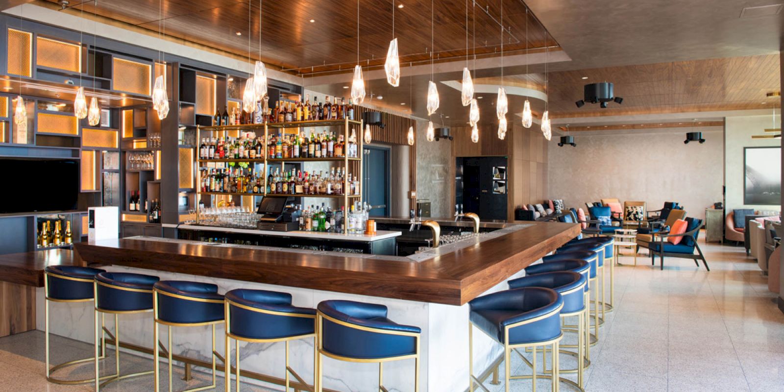 A modern hotel-style bar with a wood counter, bottle shelves, pendant lights, and stools in a lounge area.