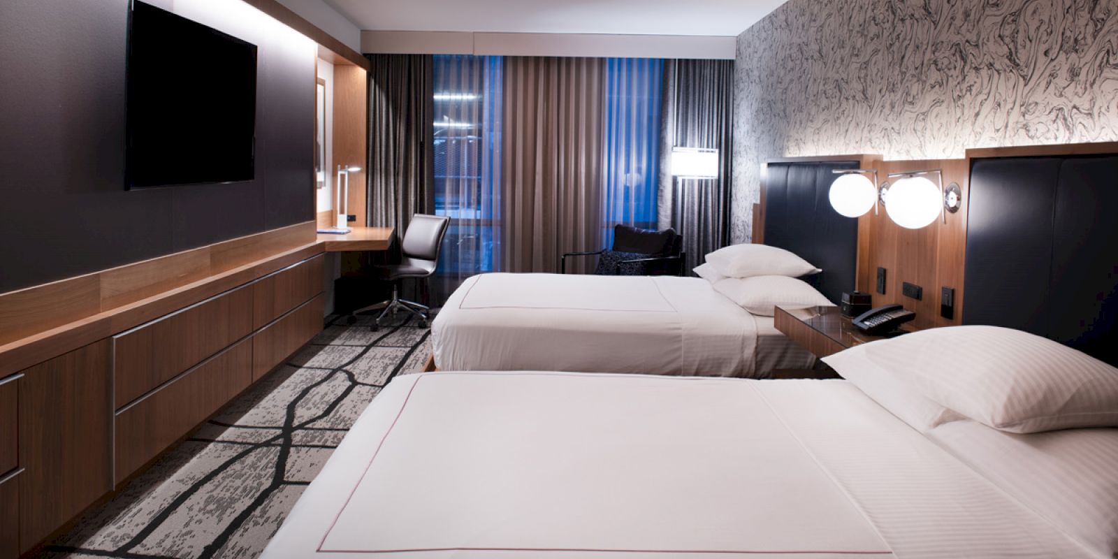 A modern hotel room with two beds, bedside lights, a wall TV, and a desk near floor-to-ceiling curtains.