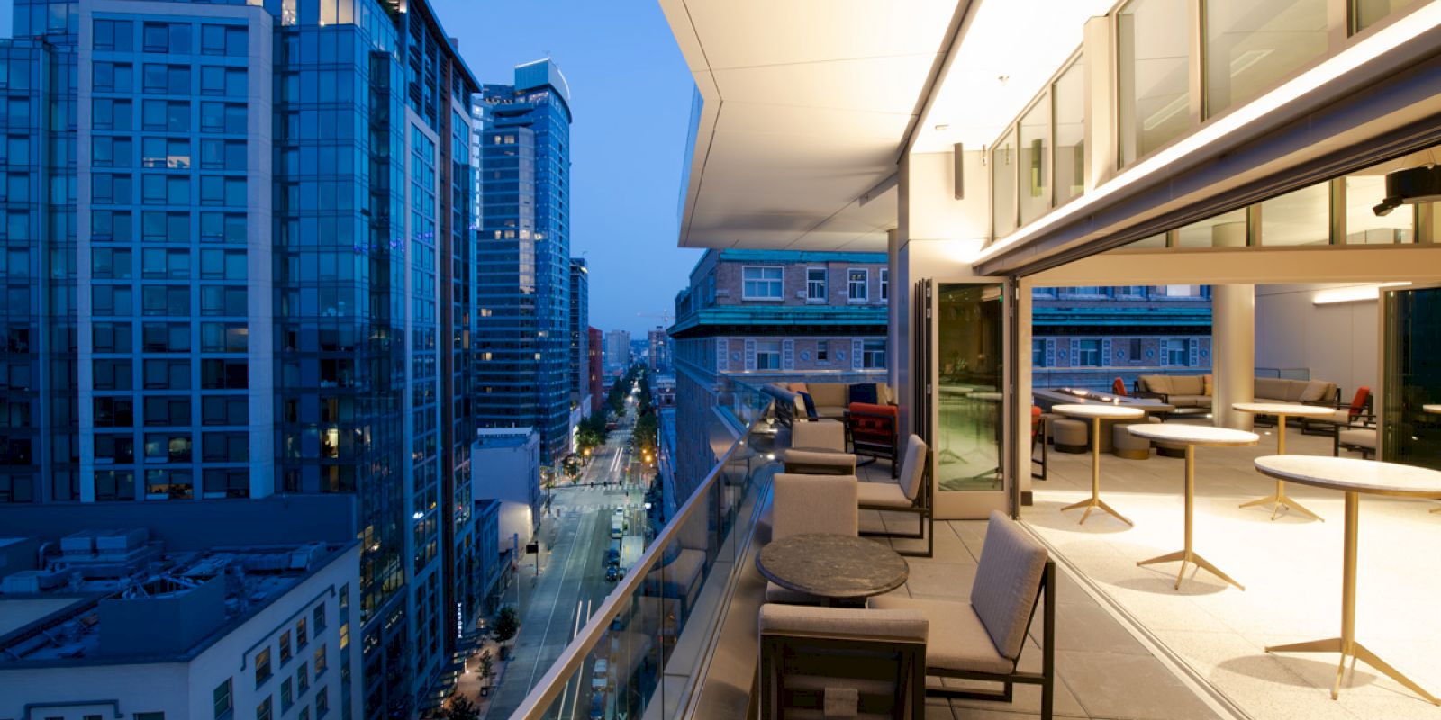 A modern high-rise rooftop terrace with lounge seating overlooks a city street lined with tall buildings.