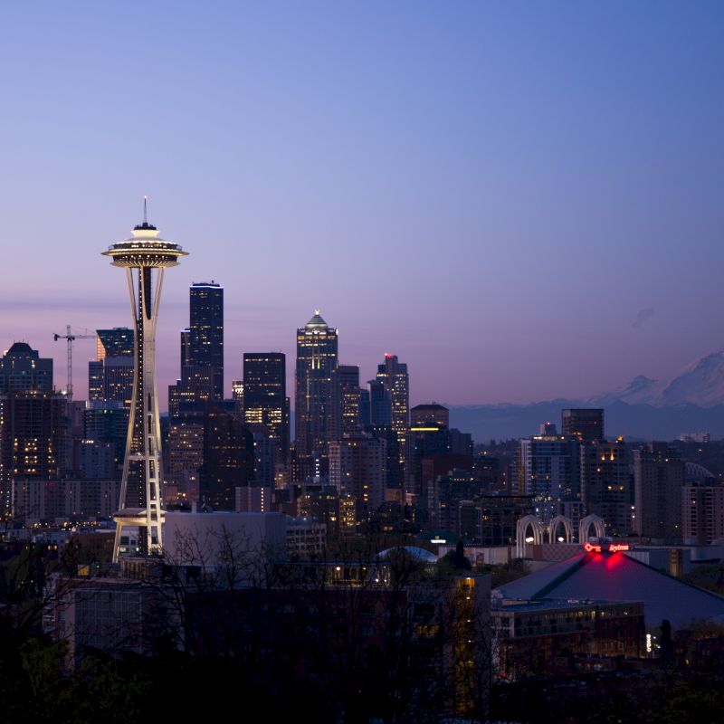 Seattle&rsquo;s Space Needle rises over a glowing city skyline at dusk, with mountains in the background.