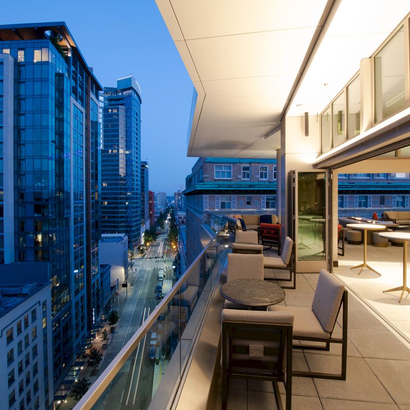 A modern high-rise rooftop terrace with lounge seating overlooks a dense city street between tall buildings at dusk.