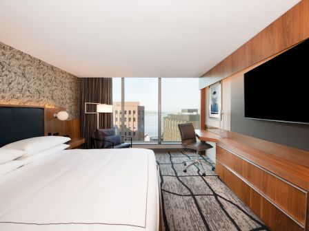 Modern hotel room with a bed, desk chair, wall TVs, wood paneling, and a city view through large windows.