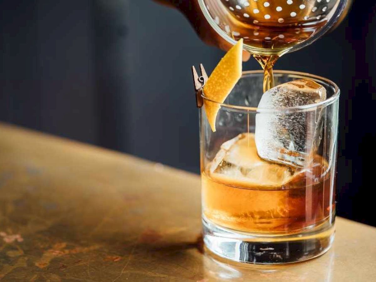 A bar pours amber liquor into an ice-filled rocks glass, garnished with a citrus wedge and swirled ice.