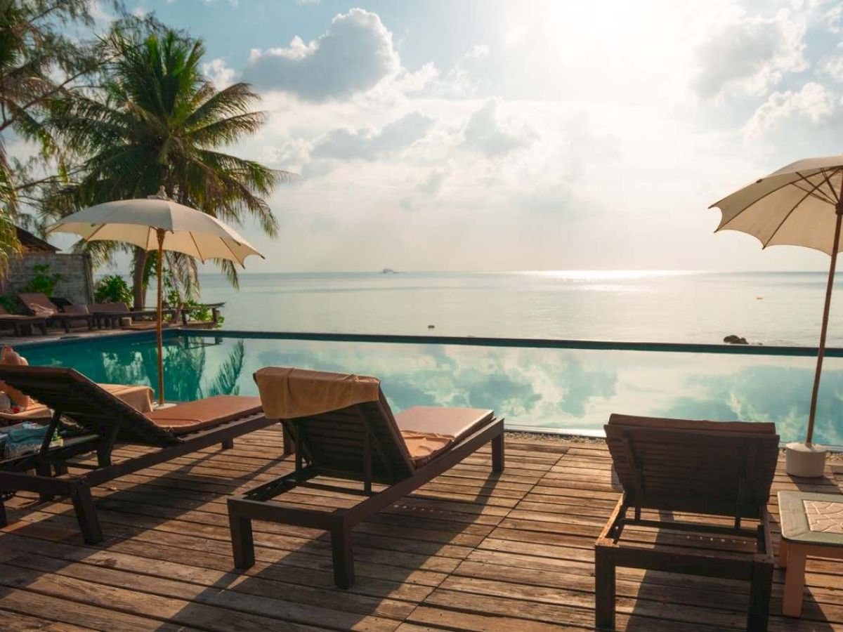 Beachfront hotel deck with lounge chairs, two umbrellas, a pool, and calm ocean under sunny clouds.