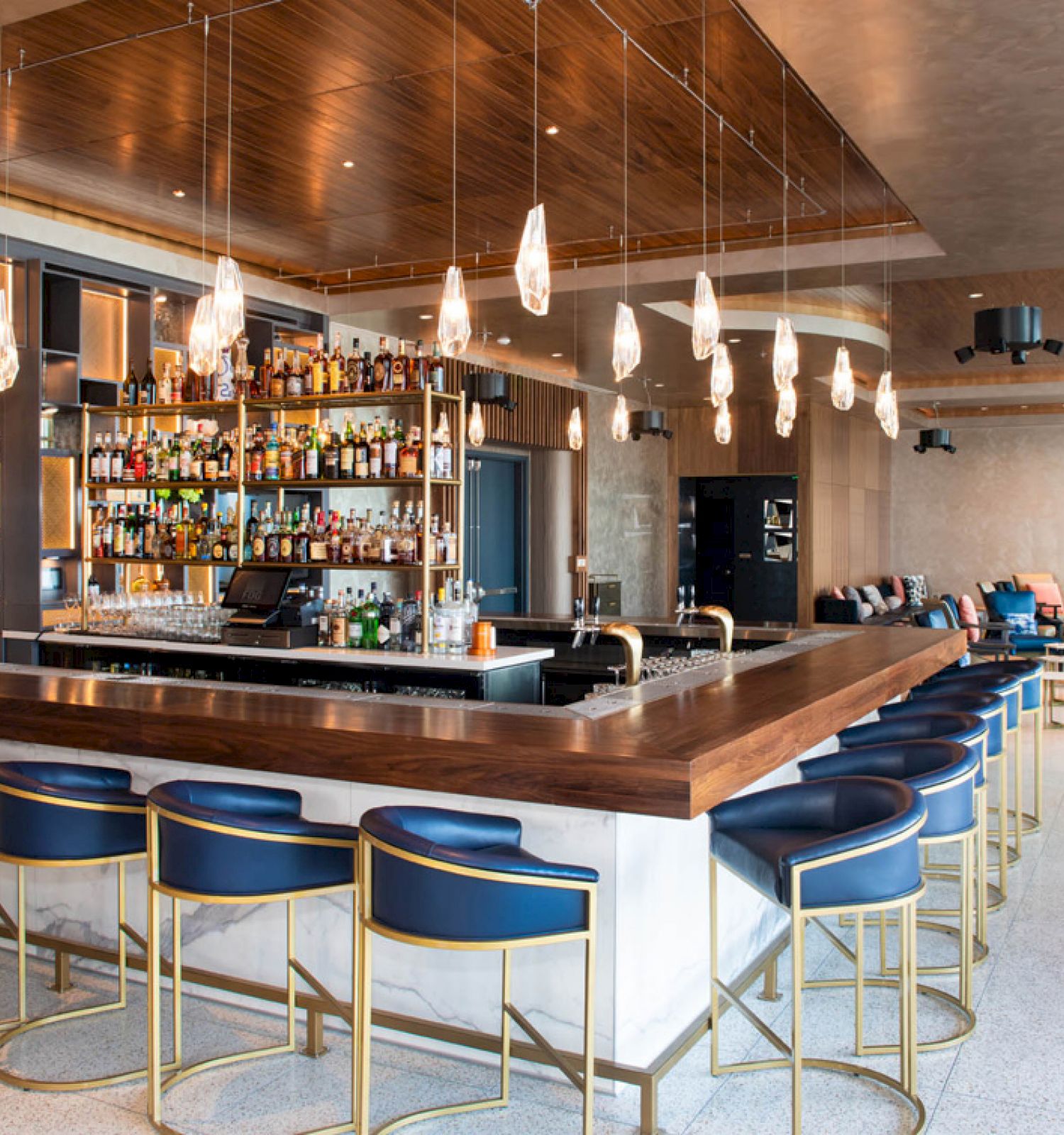 An elegant bar with a long wooden counter, stocked bottles, pendant lights, and blue leather stools in a modern lounge.