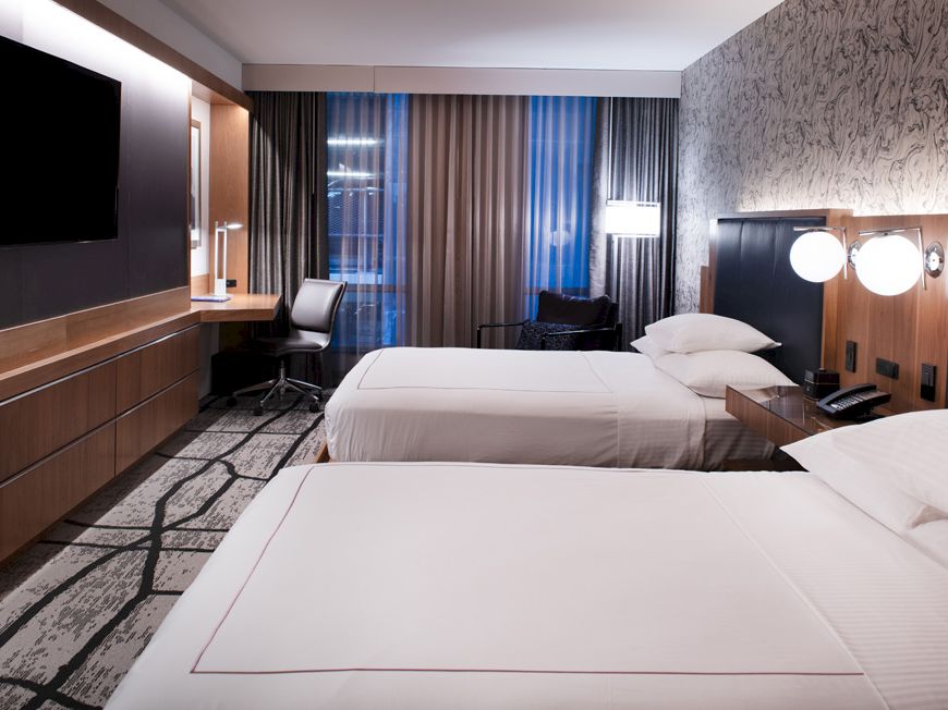A modern hotel room with two white beds, a wall TV, a desk area, and bright lamps behind dark headboards.