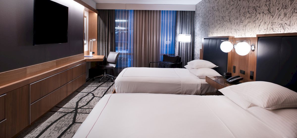 A modern hotel room with two white beds, a wall TV, a desk area, and bright lamps behind dark headboards.