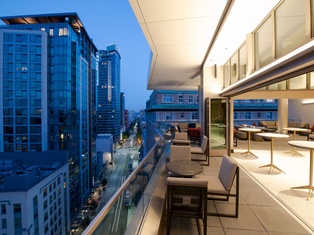 A modern rooftop terrace overlooks a city street lined with tall blue-glass skyscrapers and evening lights, from a high-rise viewpoint.
