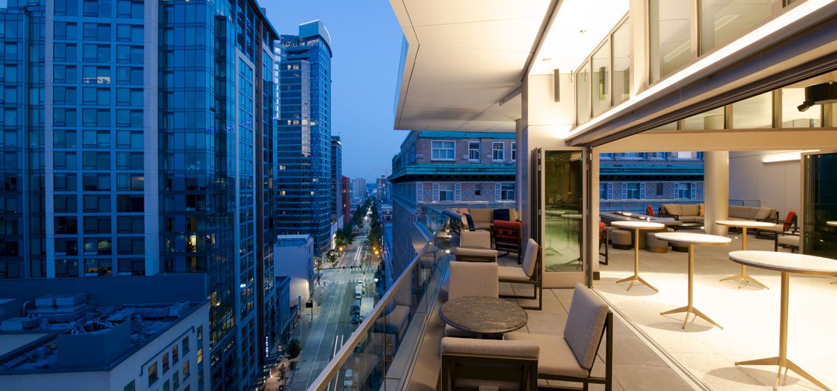A modern rooftop terrace overlooks a city street lined with tall blue-glass skyscrapers and evening lights, from a high-rise viewpoint.