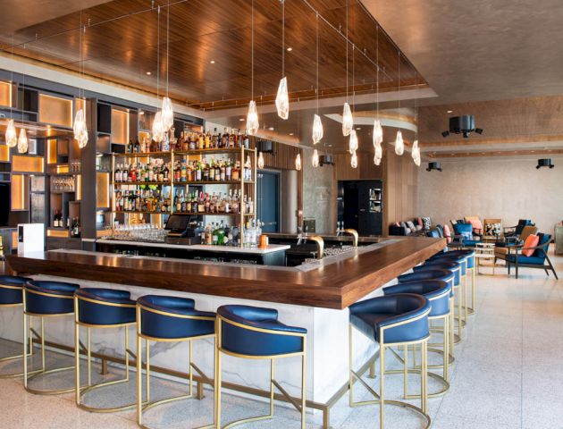 A modern hotel bar with a wood counter, backlit bottles, pendant lights, and blue stools, plus a lounge seating area.