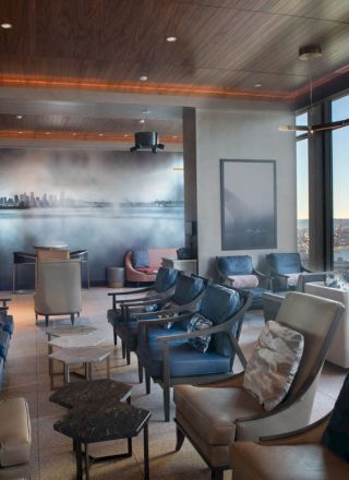 Modern lounge with cushioned armchairs and tables, panoramic windows overlooking a river and harbor at dusk.