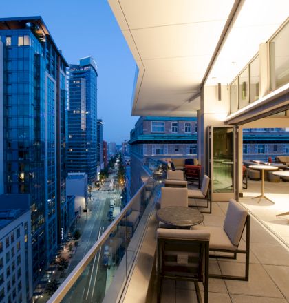 Balcony terrace with modern seating overlooks a downtown street and tall glass office buildings under a blue evening sky.