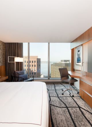 Modern hotel suite: bed, desk chair, wall-mounted TVs, wood cabinetry, patterned wallpaper, and a city view through large windows.
