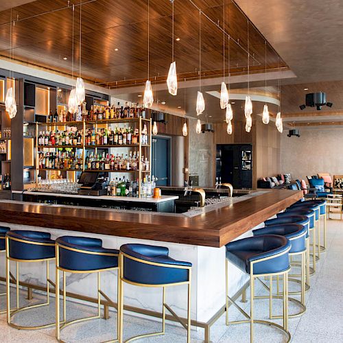 A modern hotel bar with shelves of bottles, a polished wooden counter, blue barstools, pendant lights, and a lounge seating area.
