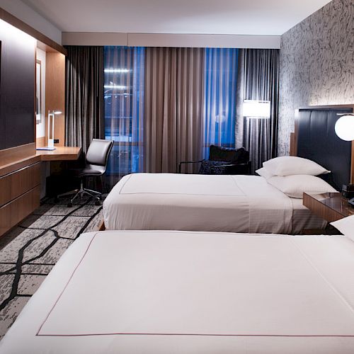 A modern hotel room with two neatly made beds, a wall TV, and a window with dark curtains.