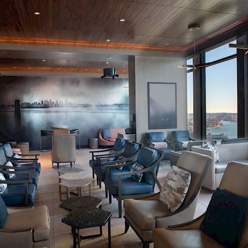A modern waterfront lounge with cushioned chairs, tables, panoramic windows, and a skyline mural wall with mist, at dusk.