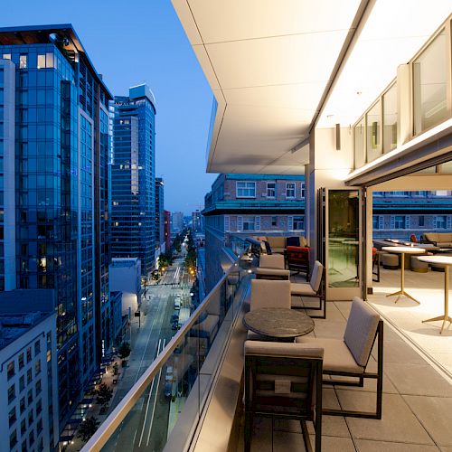 A modern rooftop terrace overlooks tall blue city skyscrapers and a street, with lounge chairs under warm light.