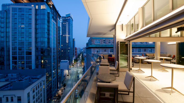 A modern high-rise rooftop lounge overlooks a city street between tall blue office buildings at dusk.
