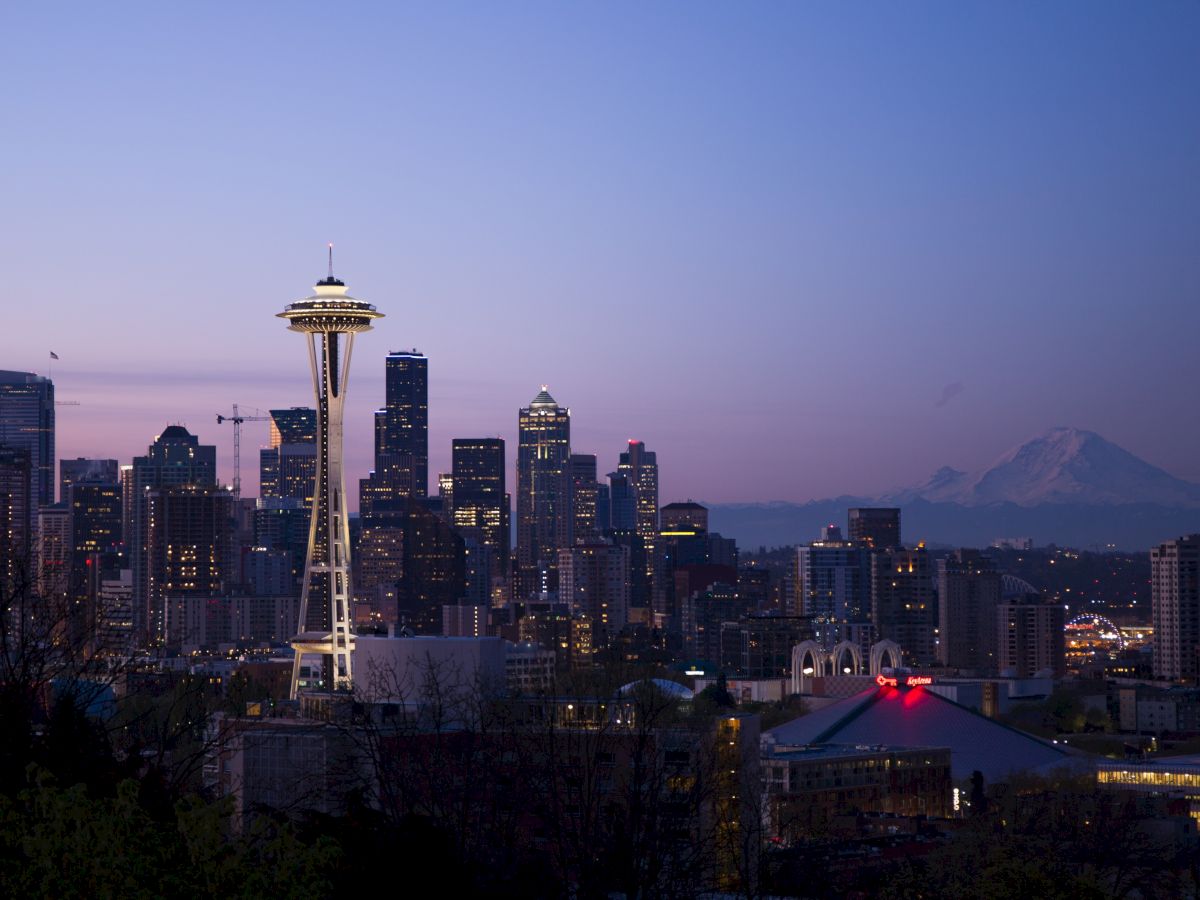 A dusk skyline of Seattle with the Space Needle glowing above downtown buildings and distant mountains.