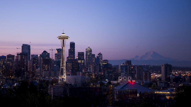 A dusk skyline of Seattle with the Space Needle glowing above downtown buildings and distant mountains.