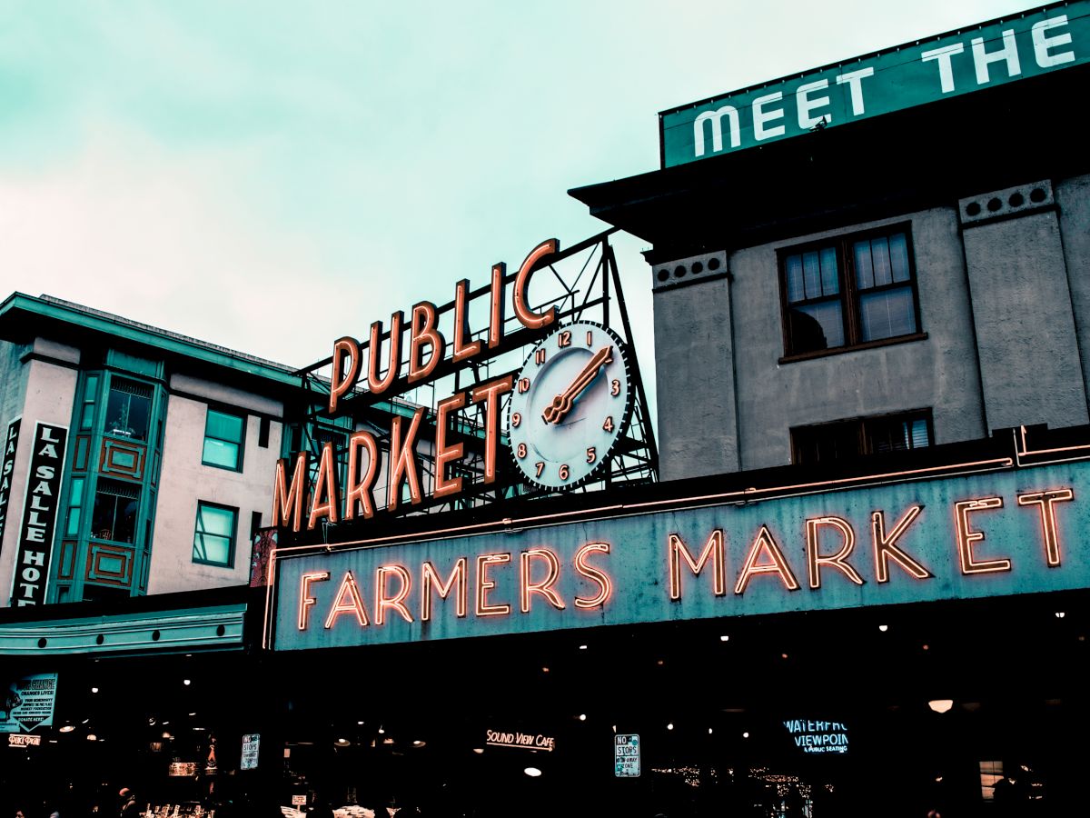 A storefront clock tower and signage reading &ldquo;Public Market&rdquo; and &ldquo;Farmers Market,&rdquo; with building fronts beneath a sky-lit dusk scene.