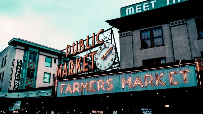 A storefront clock tower and signage reading &ldquo;Public Market&rdquo; and &ldquo;Farmers Market,&rdquo; with building fronts beneath a sky-lit dusk scene.