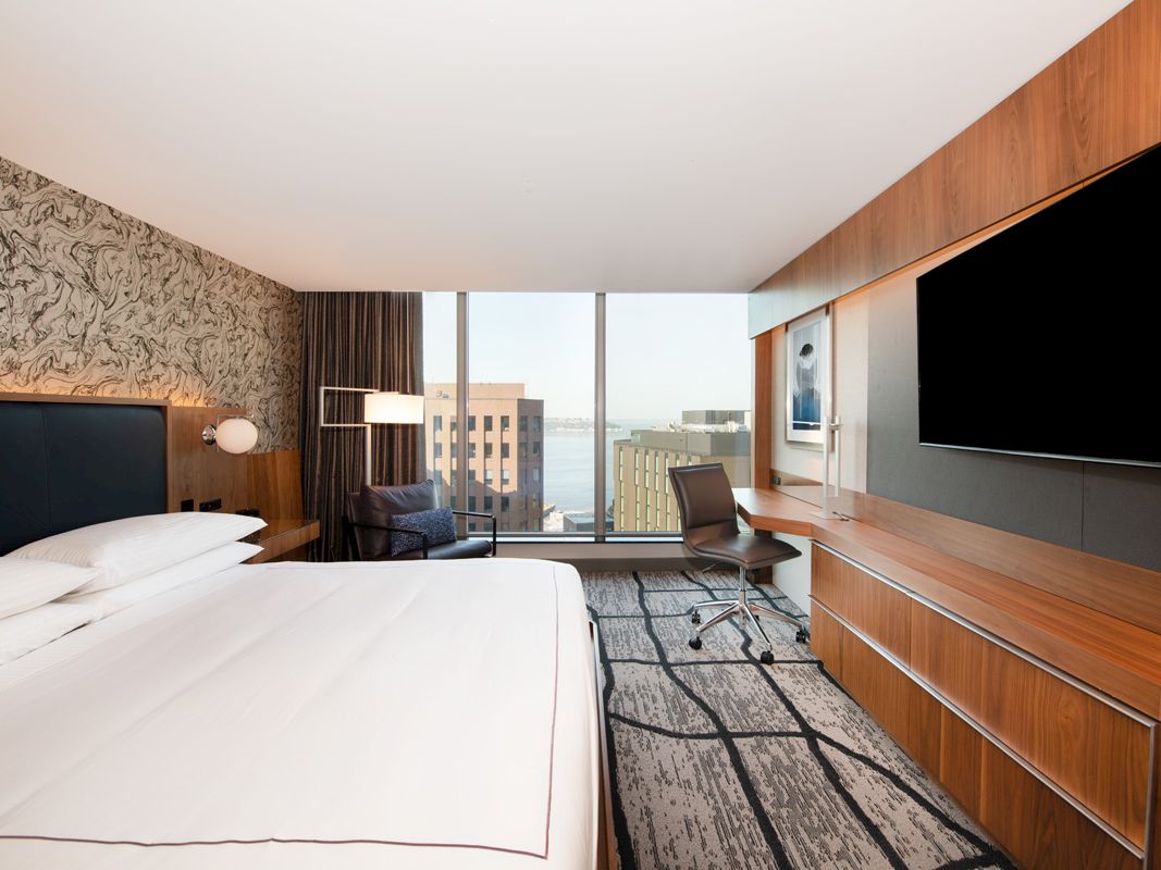 A modern hotel room with a bed, desk chair, wall-mounted TVs, and large windows overlooking the city.