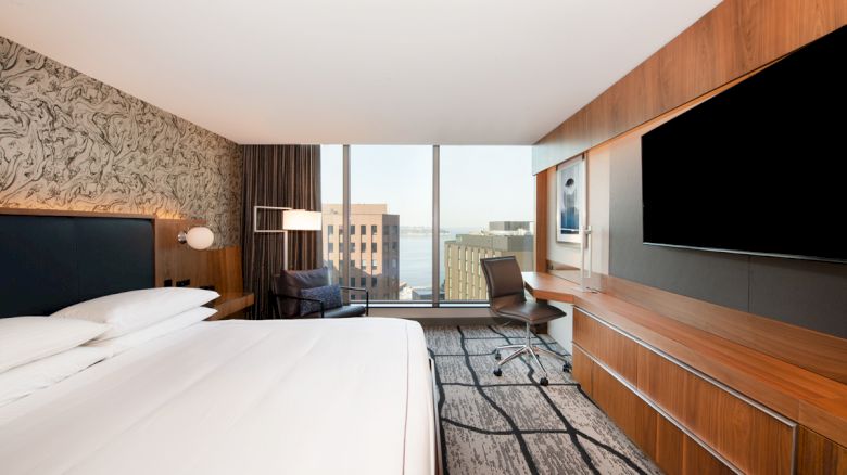 A modern hotel room with a bed, desk chair, wall-mounted TVs, and large windows overlooking the city.