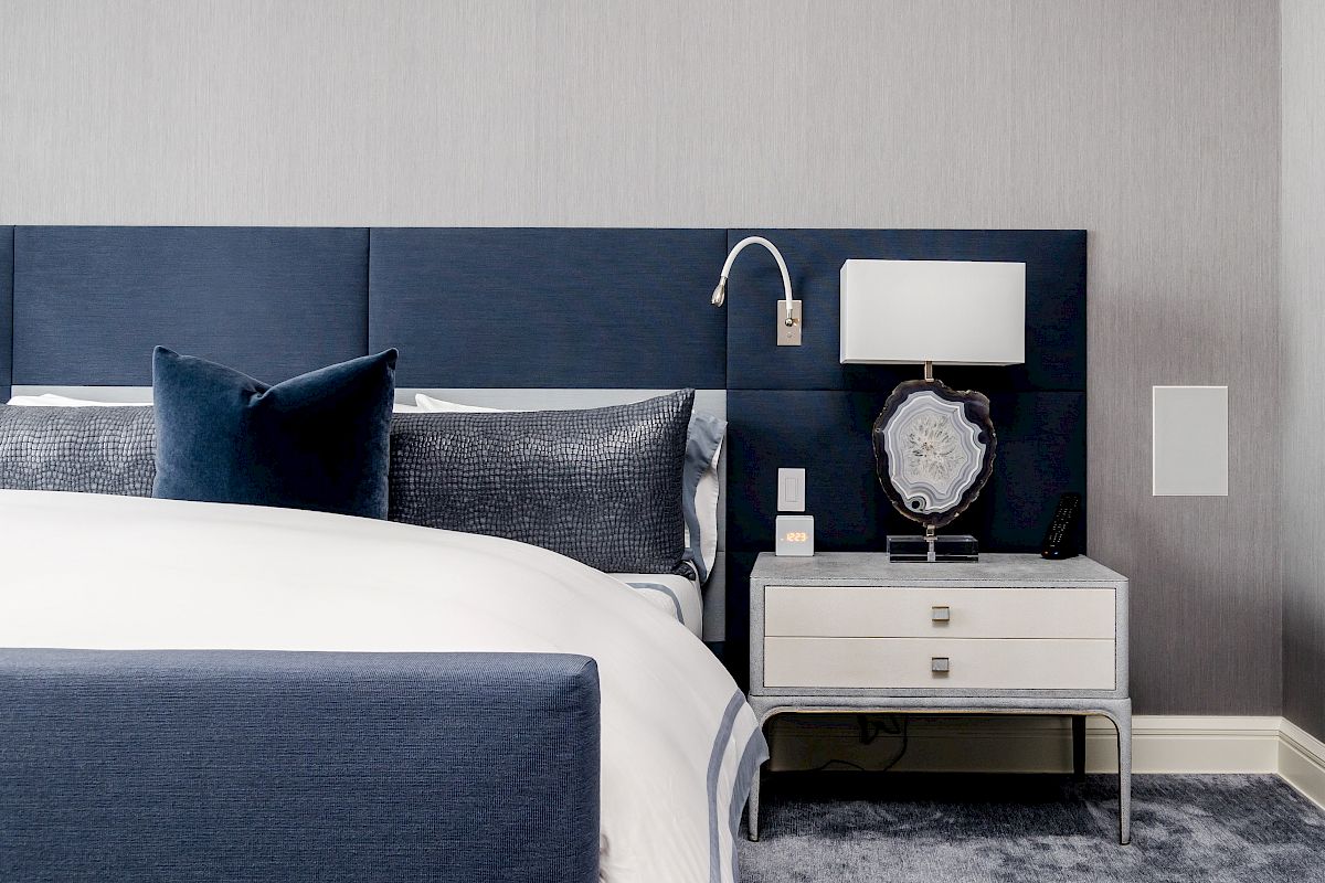 A modern bedroom with a blue bed, pillows, and a bedside table holding a lamp, clock, and decorative geode on display.