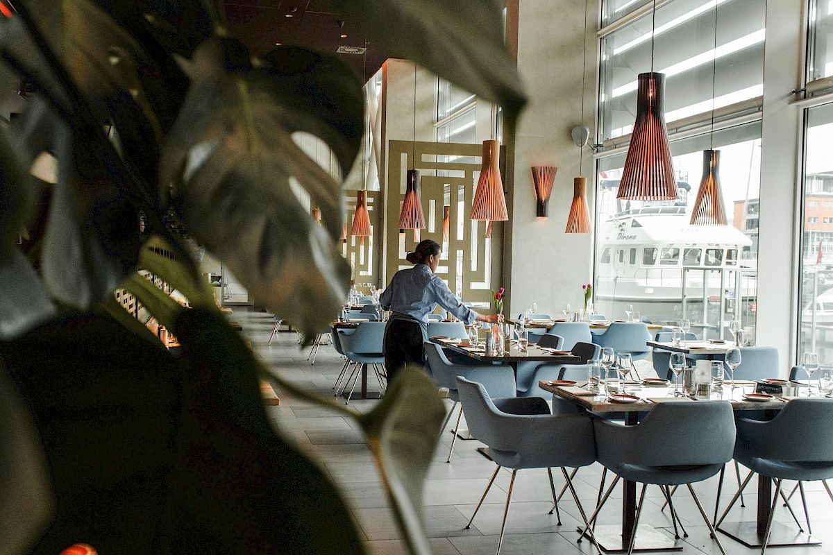 A modern restaurant dining room with blue chairs, set tables, warm pendant lights, and a person serving near big windows.