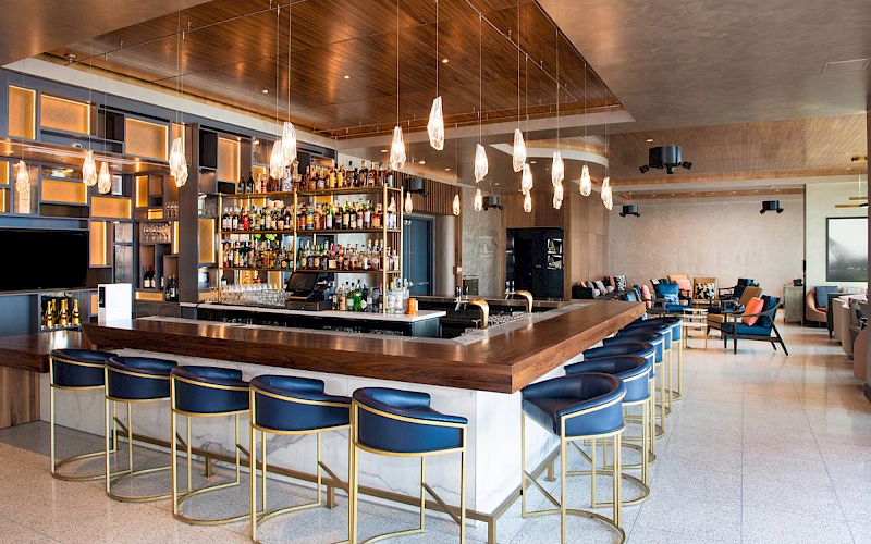 A modern hotel bar with a wooden backlit liquor shelf, pendant lights, blue stools, and lounge seating.