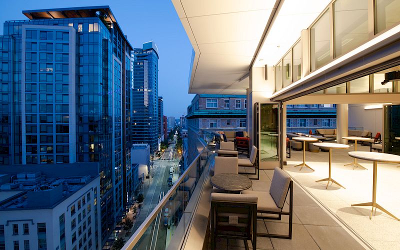 A modern high-rise rooftop terrace with lounge chairs overlooks a busy city street and glass office towers.