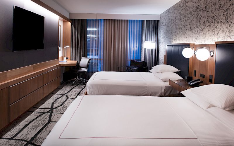 Modern hotel room with two beds, a wall-mounted TV, and warm lighting by curtained windows.