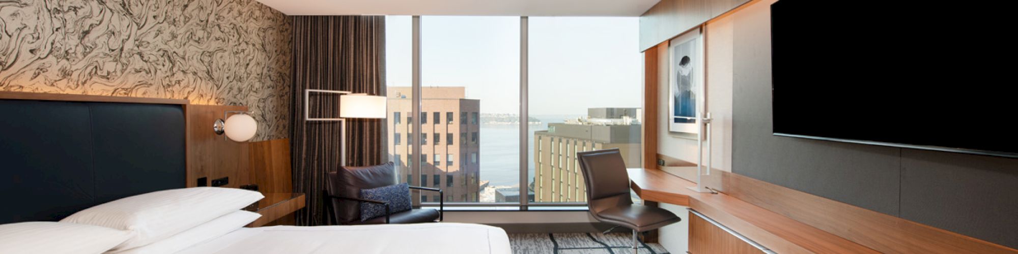 A modern hotel room with a large bed, desk chair, wall TVs, and a city view through tall windows.
