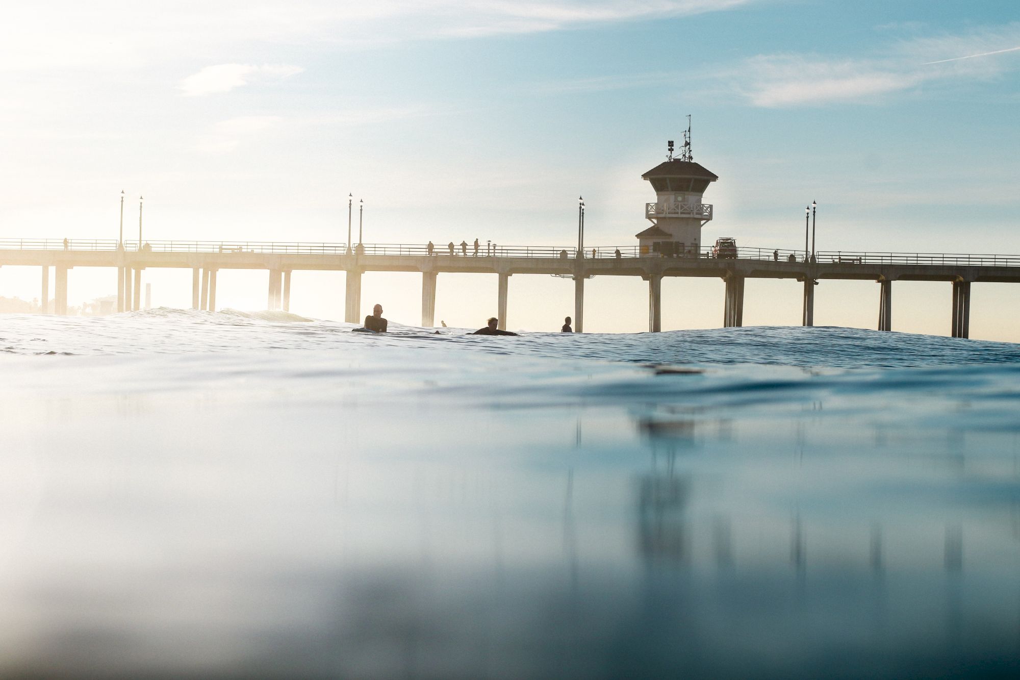 A waterfront pier with a lifeguard stand, while swimmers float in the waves under a bright, cloudy sky.